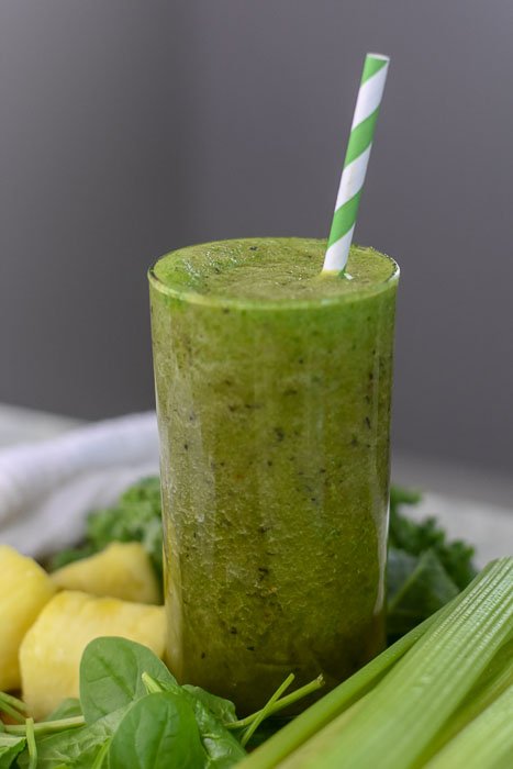 green smoothie in highball glass surrounded by fresh celery, spinach, pineapple, and kale.