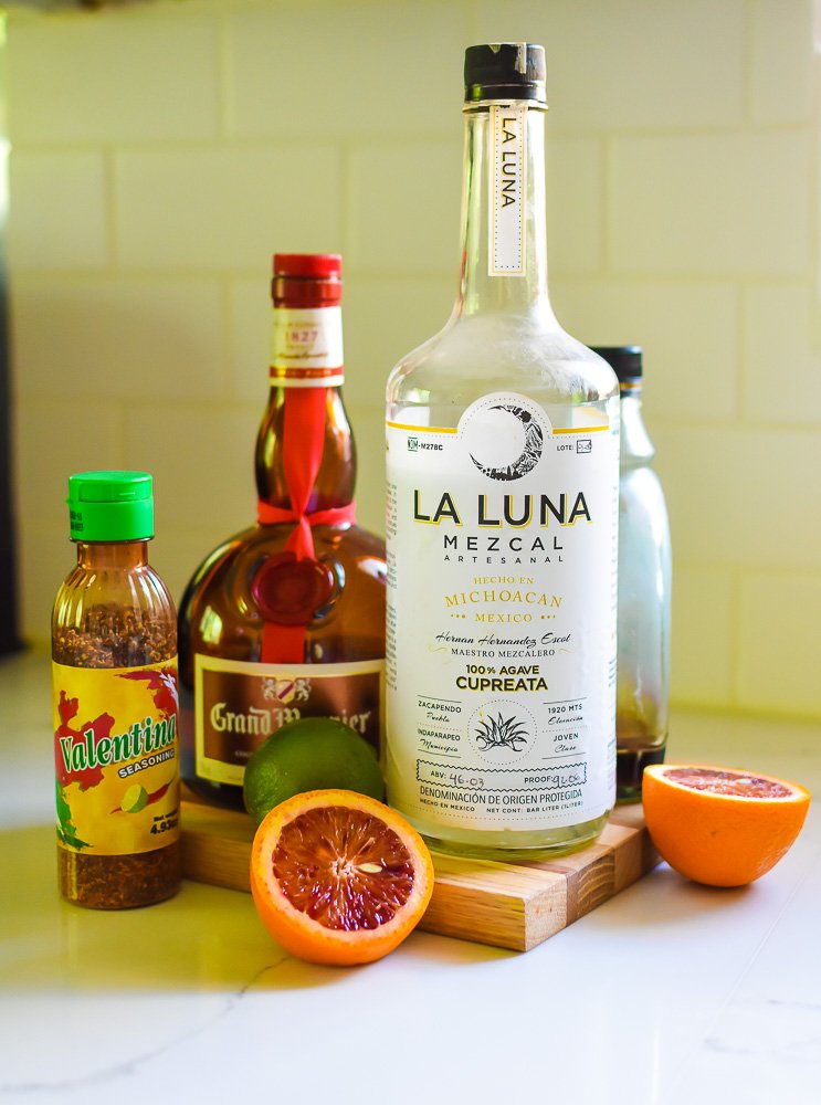 fresh lime and blood orange, bottles of mezcal, grand marnier, maple syrup, and Valentina seasoning on wooden cutting board.