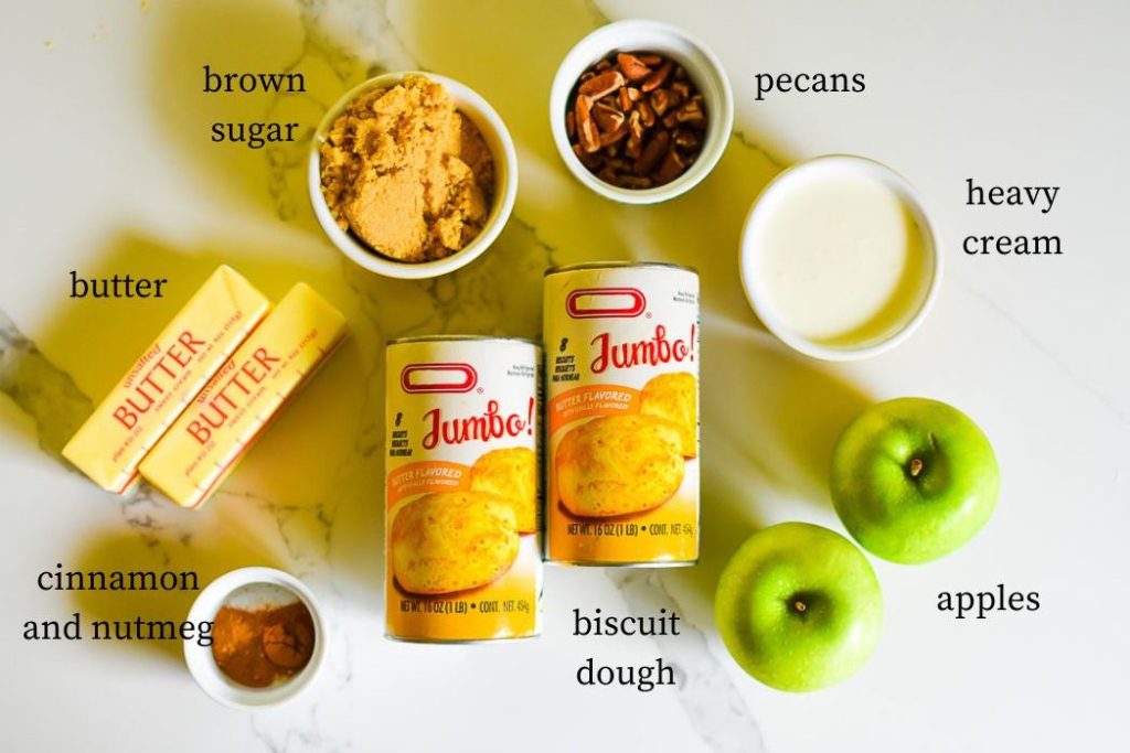 ingredients to make apple praline monkey bread loaf: brown sugar, butter, cinnamon and nutmeg, biscuit dough, pecans, heavy cream, and Granny Smith apples on countertop.