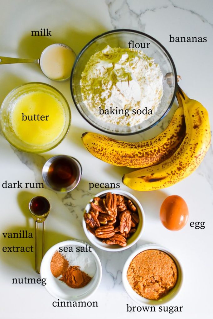 ingredients to make banana rum pecan bread on granite counter top: milk, flour, baking soda, butter, bananas, dark rum, vanilla extract, pecans, egg, sea salt, nutmeg, cinnamon, and brown sugar.