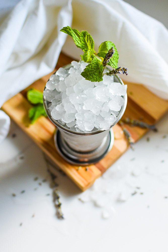 mint julep with lavender syrup in a silver cup on a wooden board surrounded by fresh mint, dried lavender, and crushed ice.