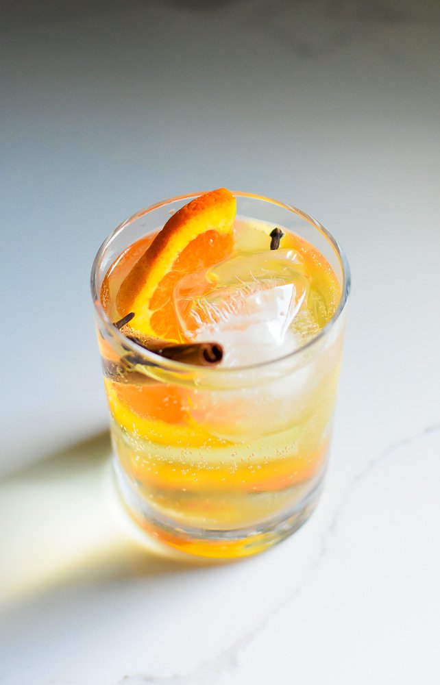 the best fall gin in tonic in a rocks glass with large ice cube, clove buds, cinnamon stick, and orange slice floating inside.