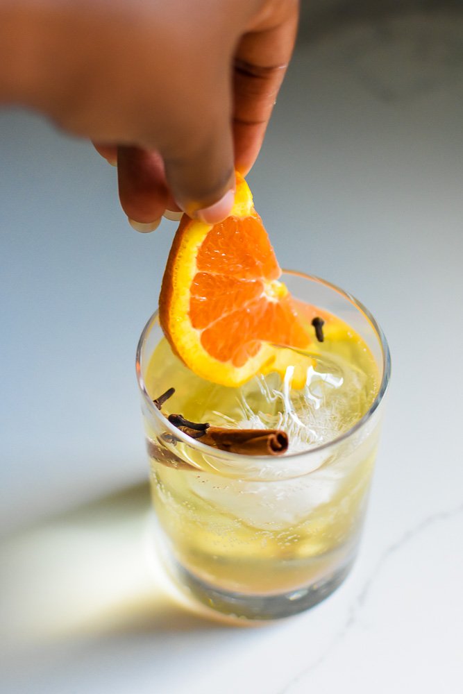 placing half an orange slice into a fall gin and tonic cocktail with cinnamon stick and clove buds.