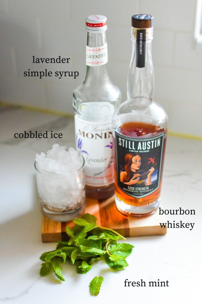 lavender mint julep drink ingredients: lavender simple syrup, bourbon whiskey, crushed ice, and fresh mint on kitchen counter top.