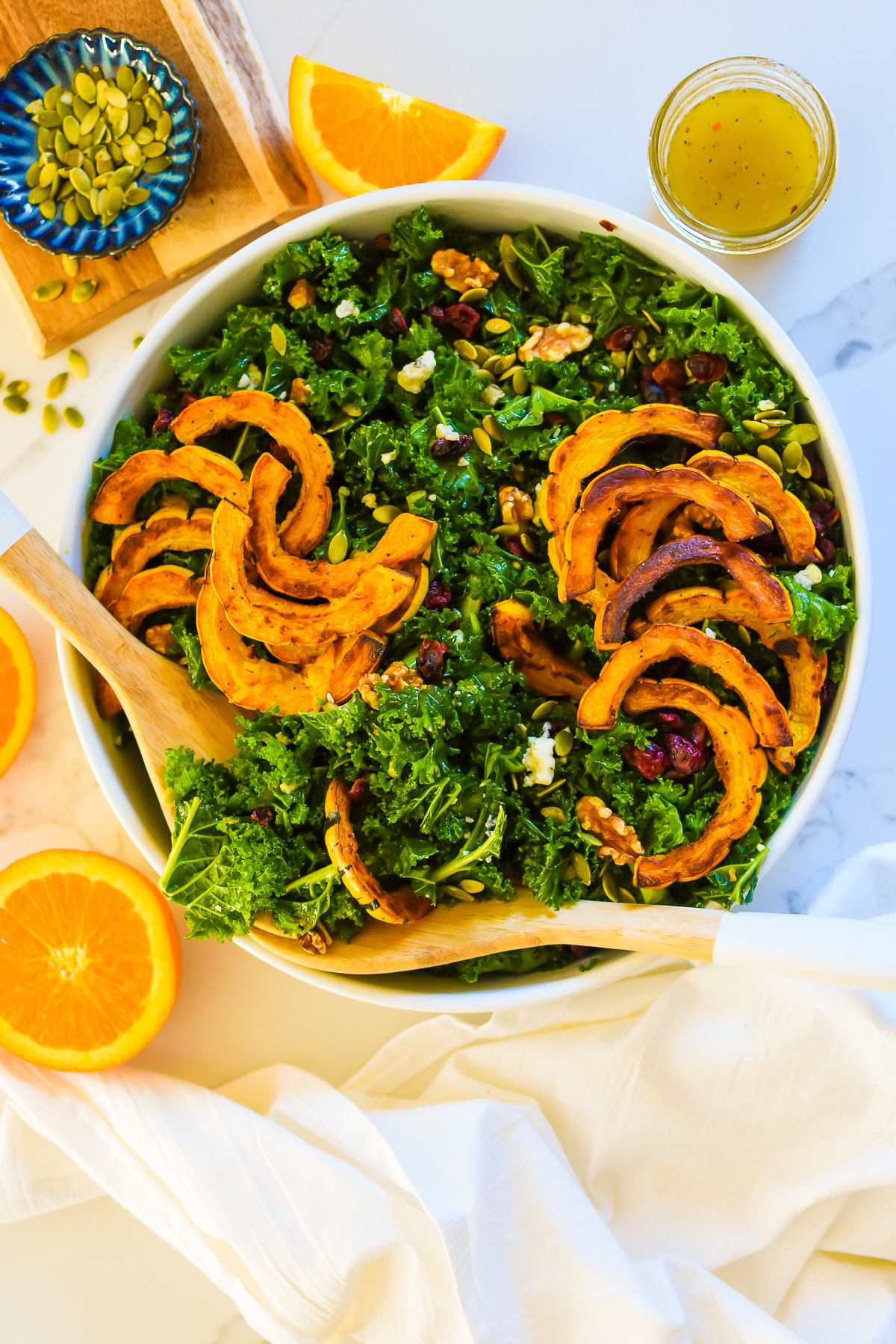 autumn harvest kale salad with roasted delicata squash, cranberries, walnuts, pumpkin seeds, and blue cheese is tossed in an orange maple vinaigrette and serve in a large white bowl perfect for Thanksgiving dinner.