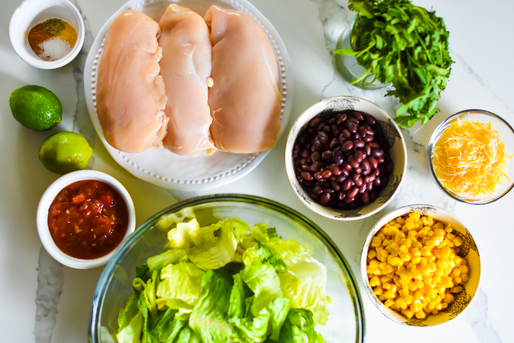 ingredients to make the best chopped southwestern chicken salad on granite counter top: spices, boneless skinless chicken breast, limes, salsa, chopped romaine, black beans, corn, cilantro, and monterrey jack cheese.