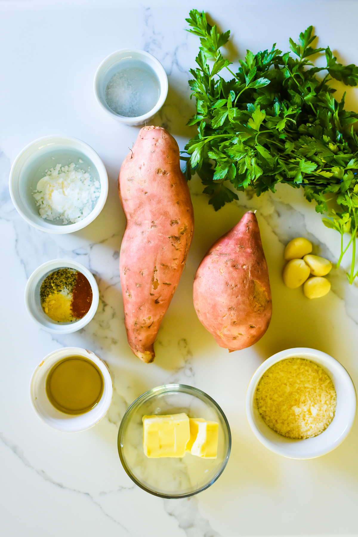 ingredients for crispy oven baked sweet potato fries on granite counter top: sweet potatoes, parsley, sea salt, cornstarch, spices, avocado oil, butter, parmesan cheese, and garlic cloves.