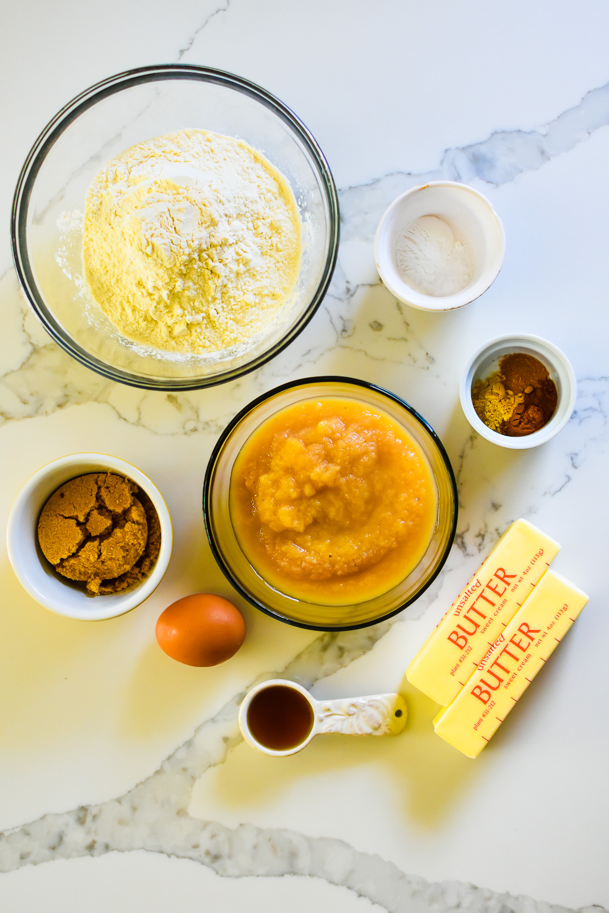 ingredients to make the best applesauce bread on granite counter top: flour, baking powder, spices, applesauce, brown sugar, egg, butter, and maple syrup.