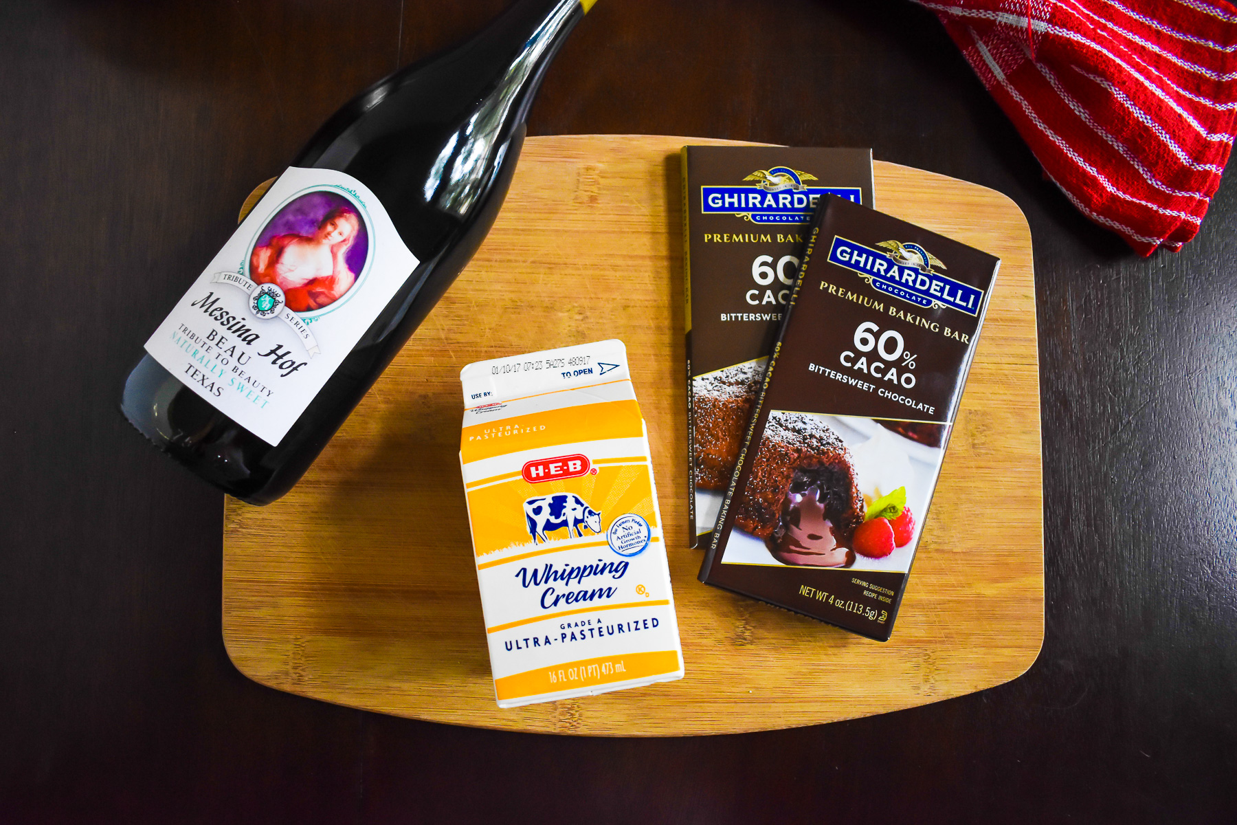 bottle of red wine, carton of whipping cream and bars of bittersweet chocolate on wooden cutting board.