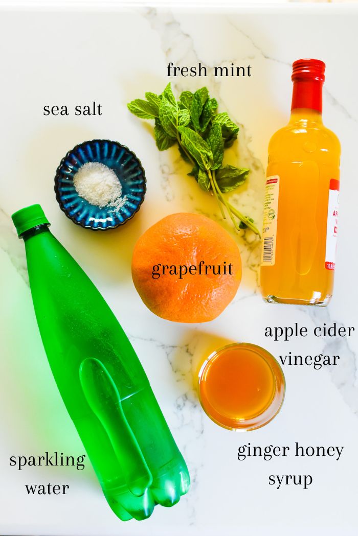 ingredients to make a sparkling grapefruit mint mocktail on counter top, including fresh mint, ruby red grapefruit, apple cider vinegar, sea salt, sparkling water, and ginger honey syrup.