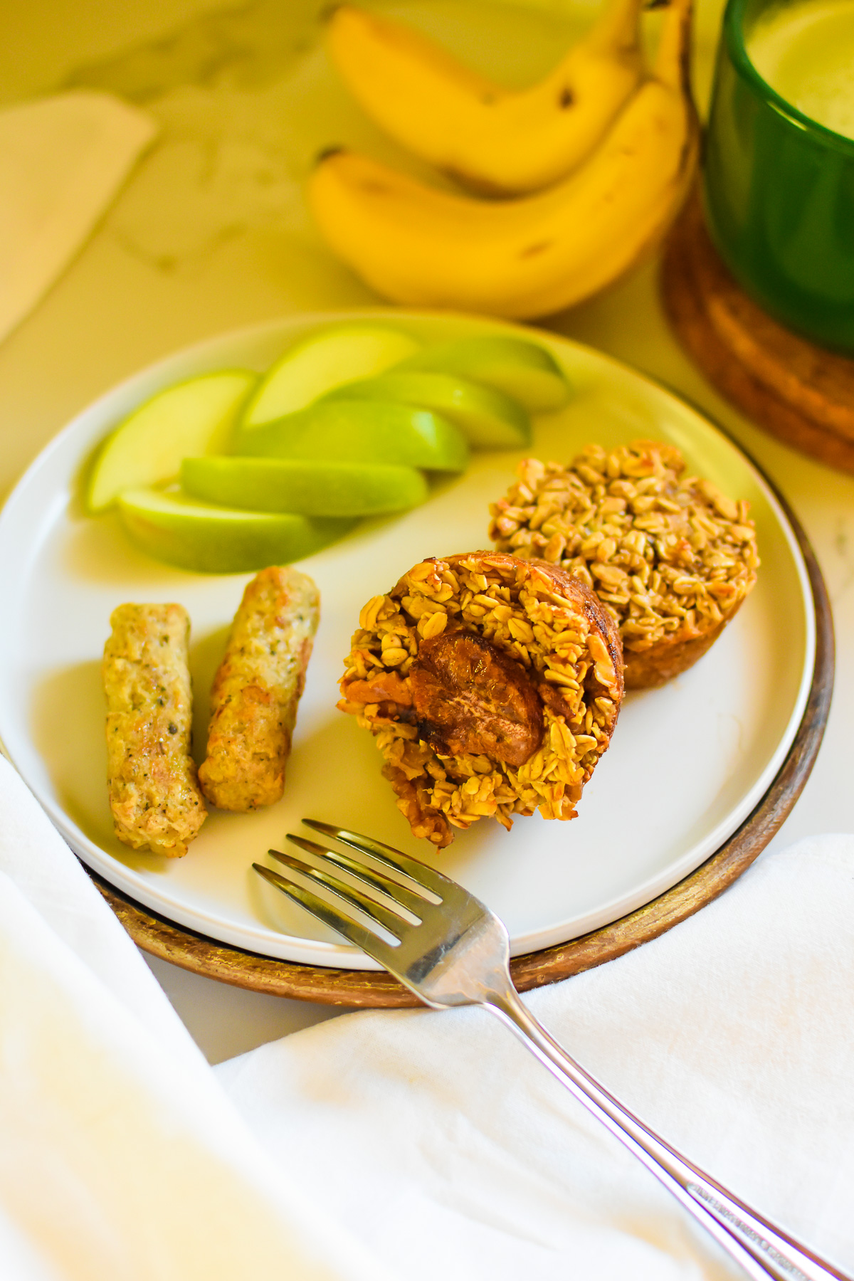 the best banana bread baked oatmeal muffins made easy in just 30 minutes served on a white plate with slices of green apple and two breakfast sausage links.