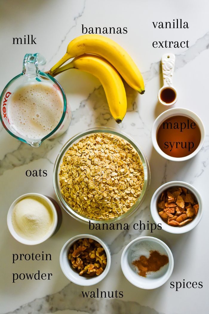 ingredients to make banana baked protein oatmeal from scratch on granite countertop, including fresh bananas, milk, vanilla extract, maple syrup, rolled oats, protein powder, banana chips, walnuts, and sweet warming spices.