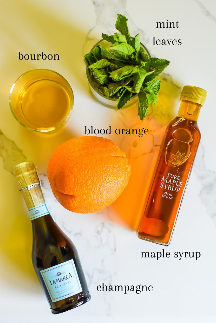 ingredients to make the best blood orange bourbon smash at home on counter top, including sparkling wine, blood orange, bourbon, fresh mint, and maple syrup.