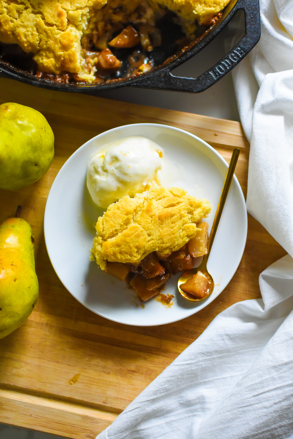 the best pear cobbler made with spiced pears and biscuit topping served on a plate with a scoop of honey vanilla ice cream next to fresh pears on a wooden cutting board.