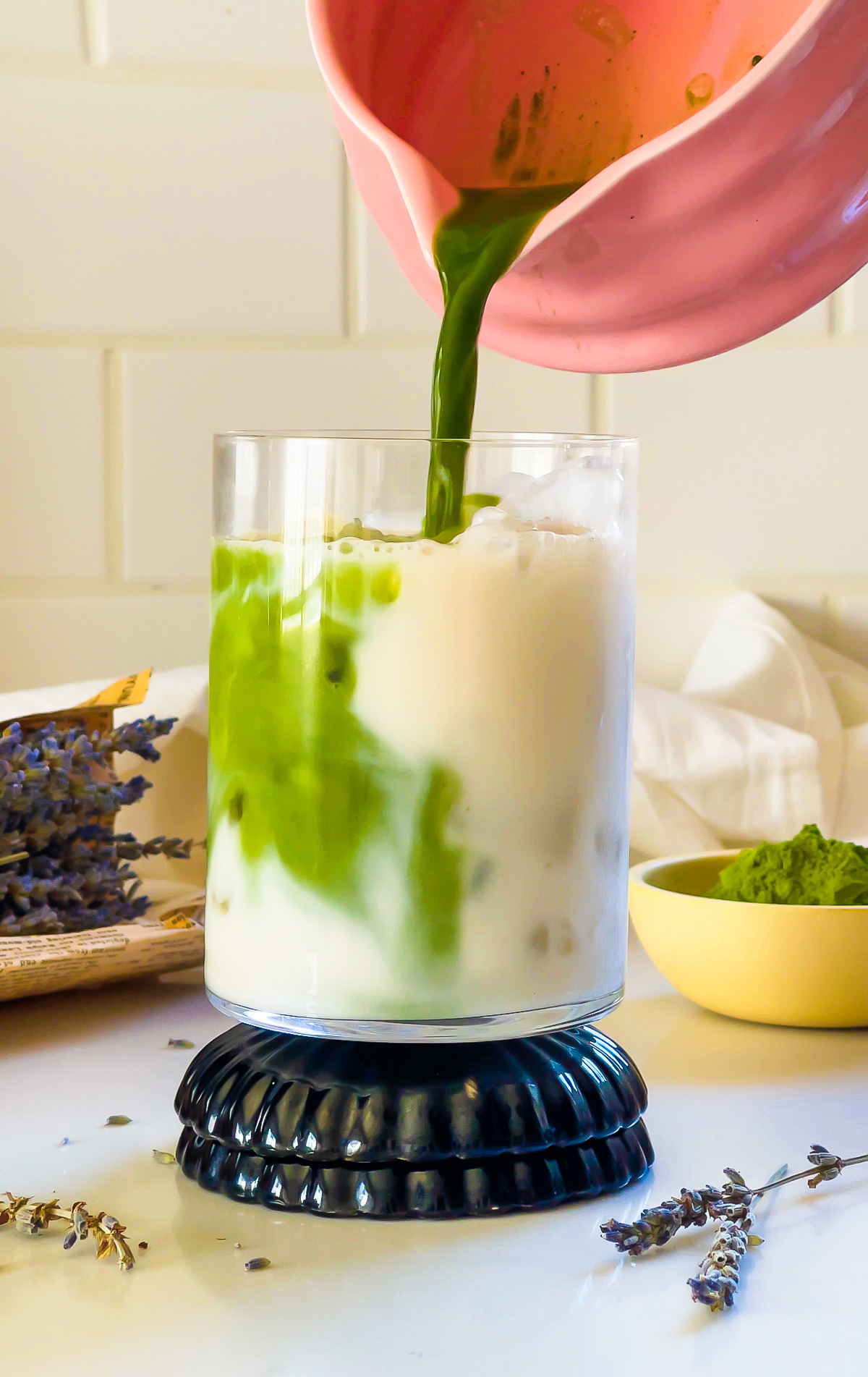 pouring lavender flavored matcha tea into glass of iced milk on stack of blue coasters.
