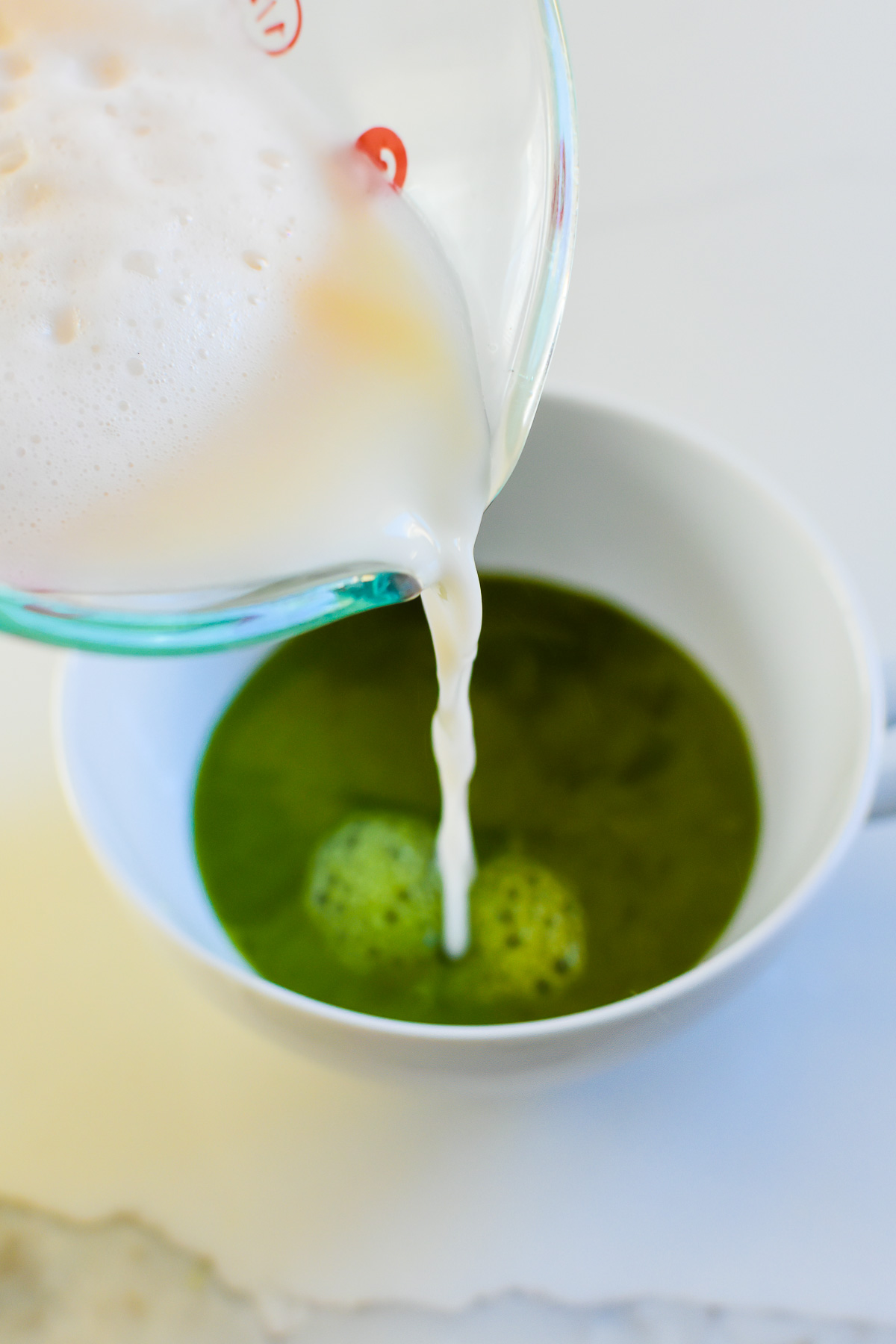 pouring foamy steamed milk into tea cup of bright green matcha green tea.
