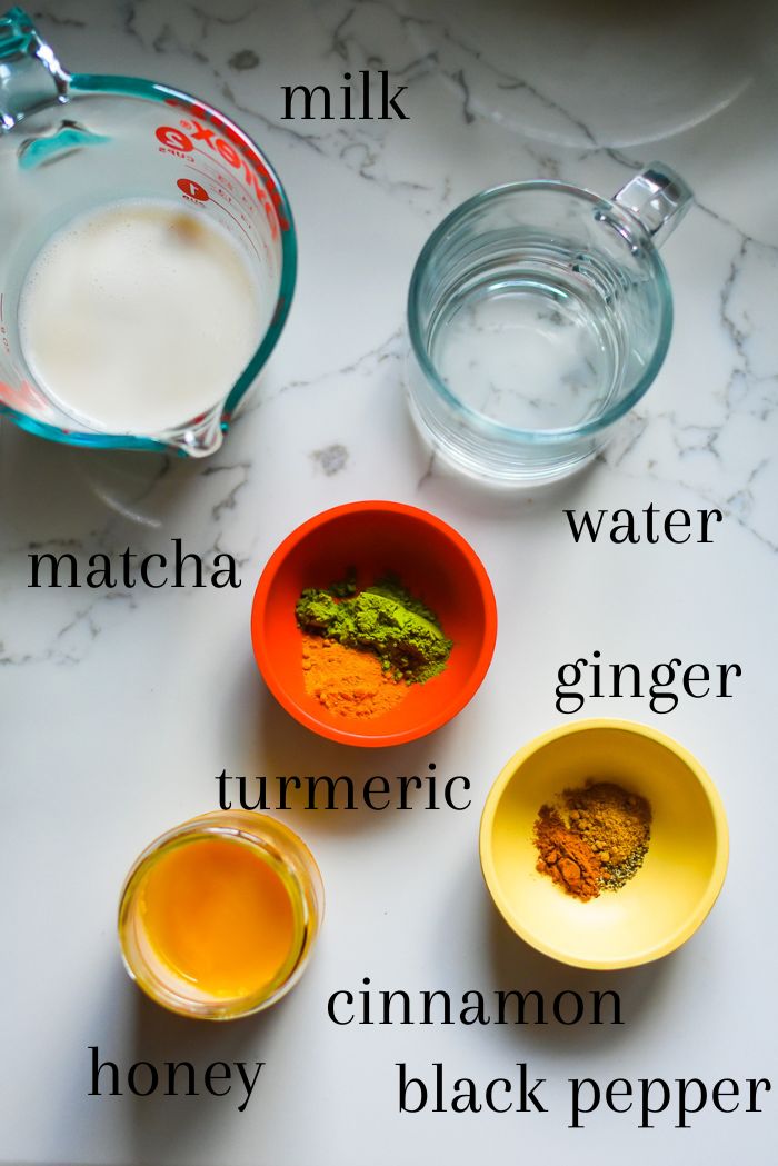 ingredients to make a homemade golden matcha latte on granite counter top, including milk, water, organic ceremonial grade matcha powder, ground turmeric, ginger, cinnamon, black pepper, and honey.