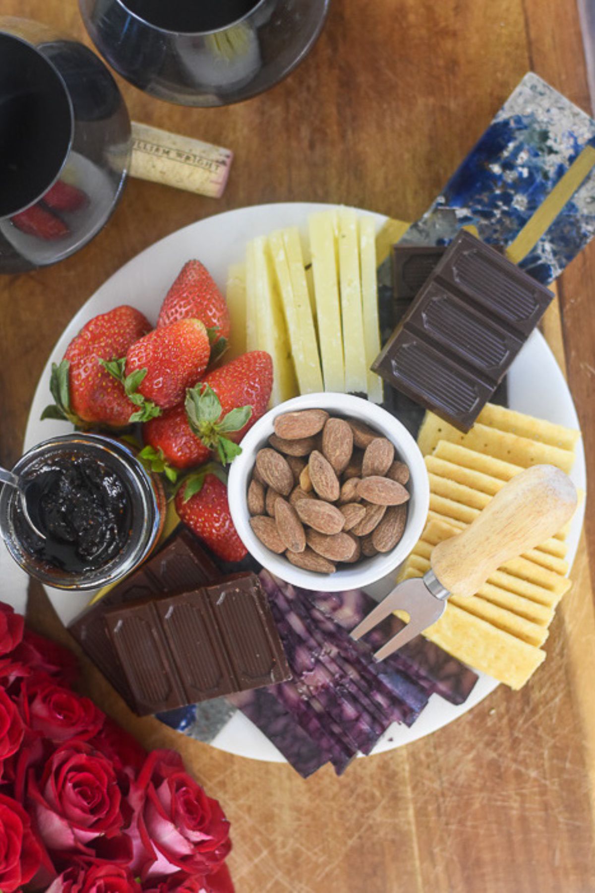 The perfect cheese board for two made with fresh berries, nuts, cheeses, crackers, chocolate, and preserves and served with red wine on a round marble platter.