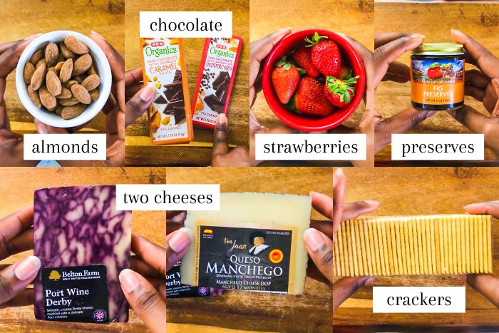 ingredients to make a date night cheese platter for two at home on wooden cutting board: almonds, chocolate bars, fresh strawberries, jar of fig preserves, port wine derby cheese, queso manchego, and buttery crackers.