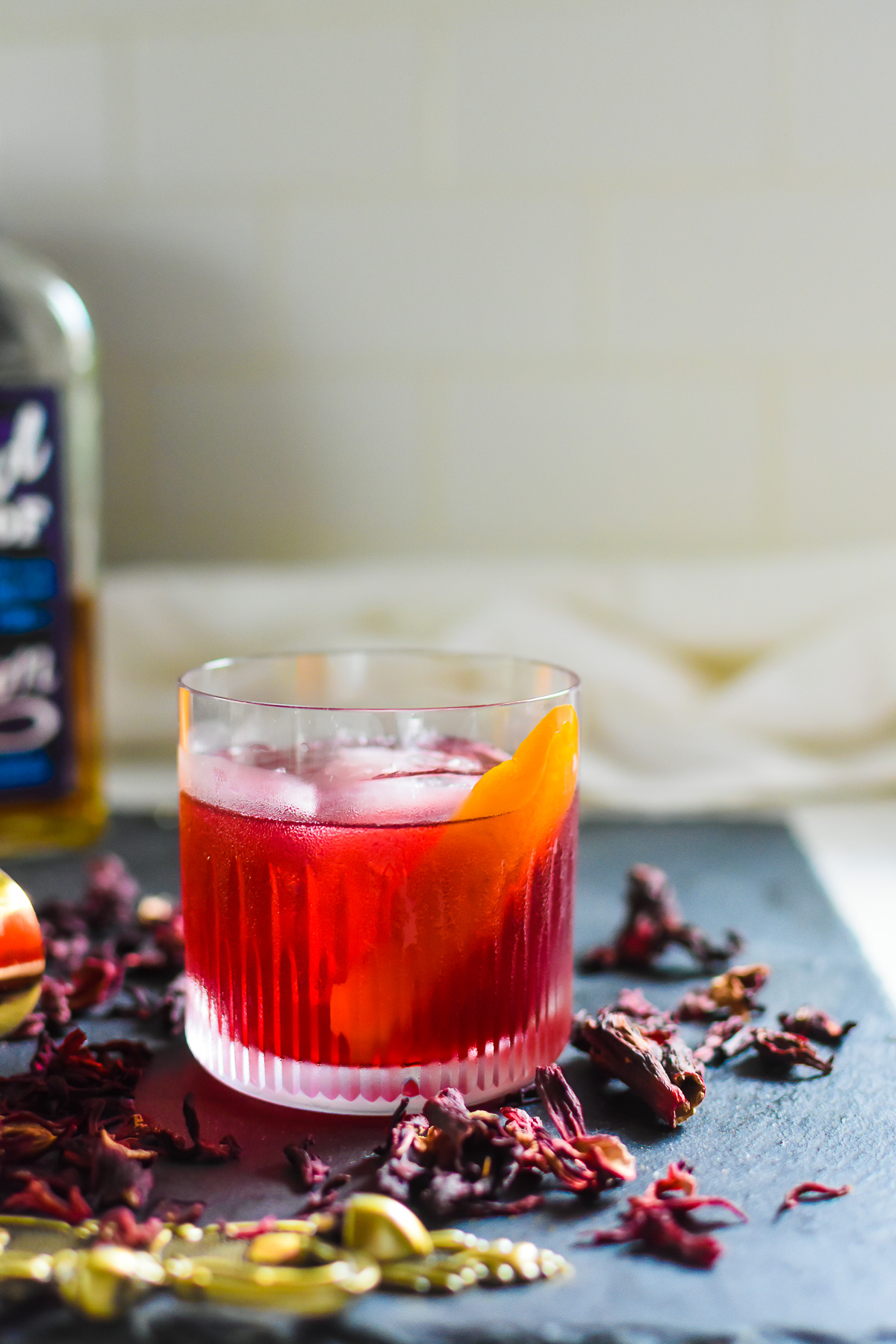 homemade hibiscus old fashioned cocktail on slate board surrounded by dried hibiscus, zobo or jamaíca flowers.