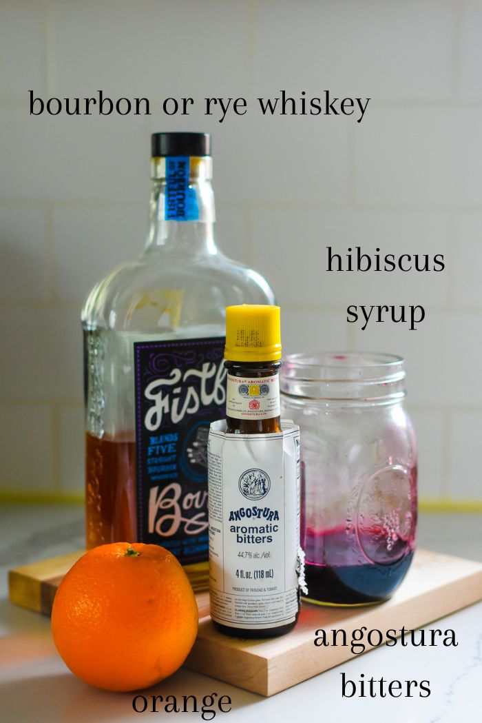Ingredients to make a quality hibiscus old fashioned cocktail at home on counter top, including bottles of bourbon whiskey, angostura bitters, and homemade hibiscus syrup and an orange.