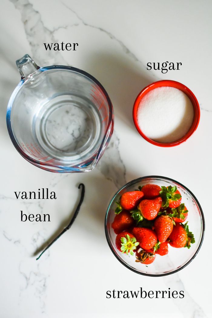Ingredients to make strawberry vanilla simple syrup at home on granite counter top, including a bowl of fresh strawberries, one vanilla bean, dish of sugar, and water.