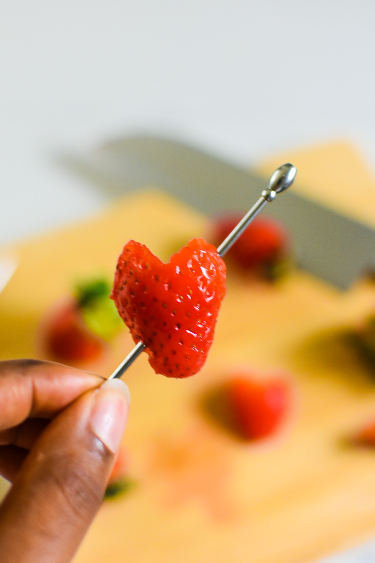 brown hand holding halved strawberry cut in a heart shape on a cocktail pick.
