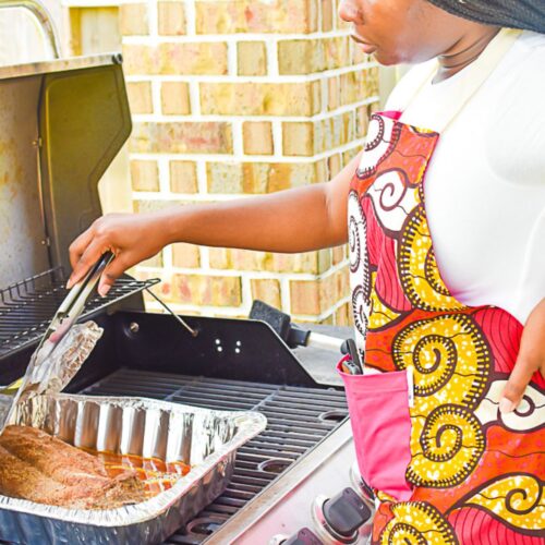 Jazzmine tending to rack of baby back ribs on a gas grill while wearing a colorful ankara apron.