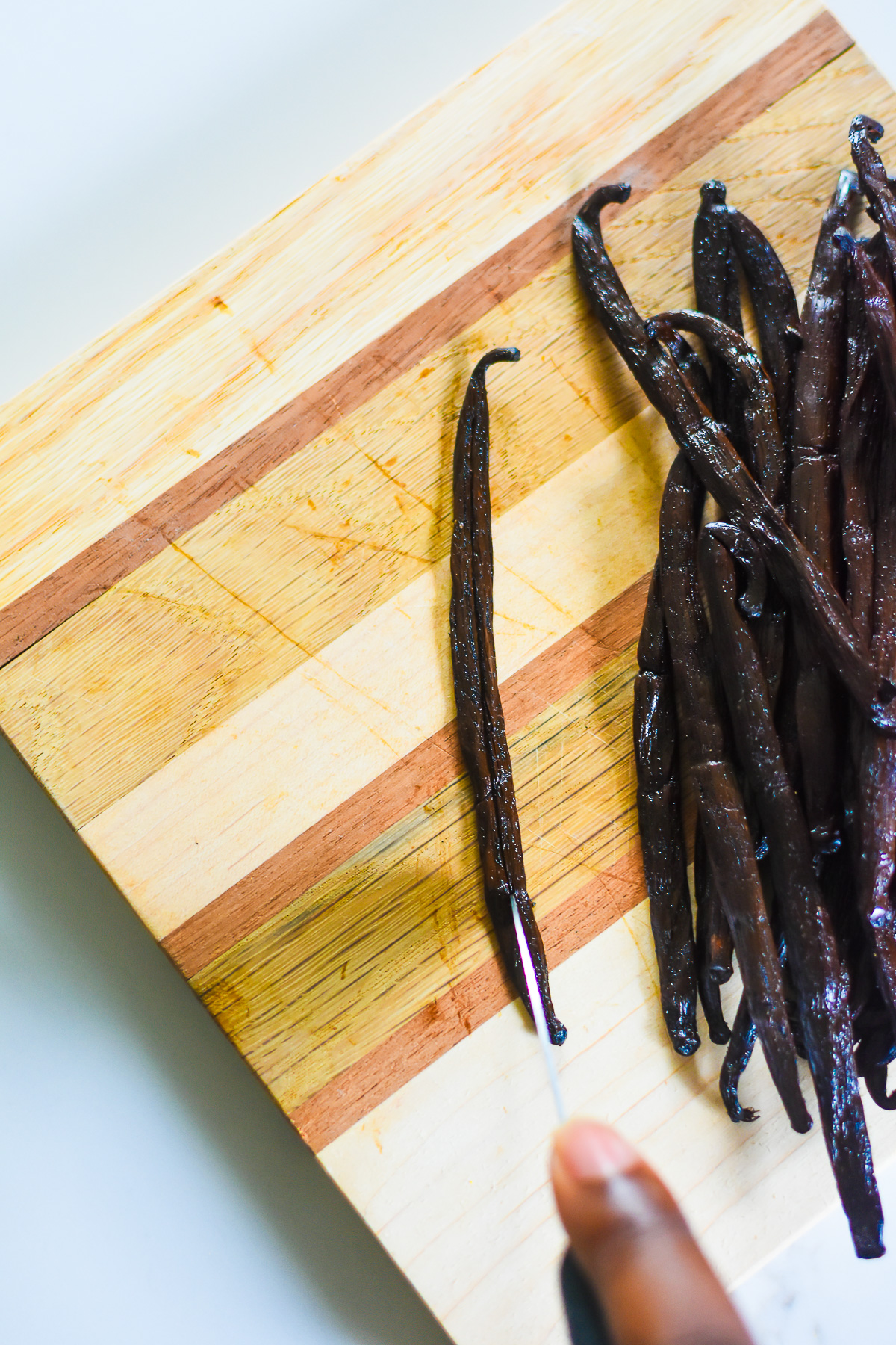 brown hand splitting a vanilla bean with a knife on a cutting board.