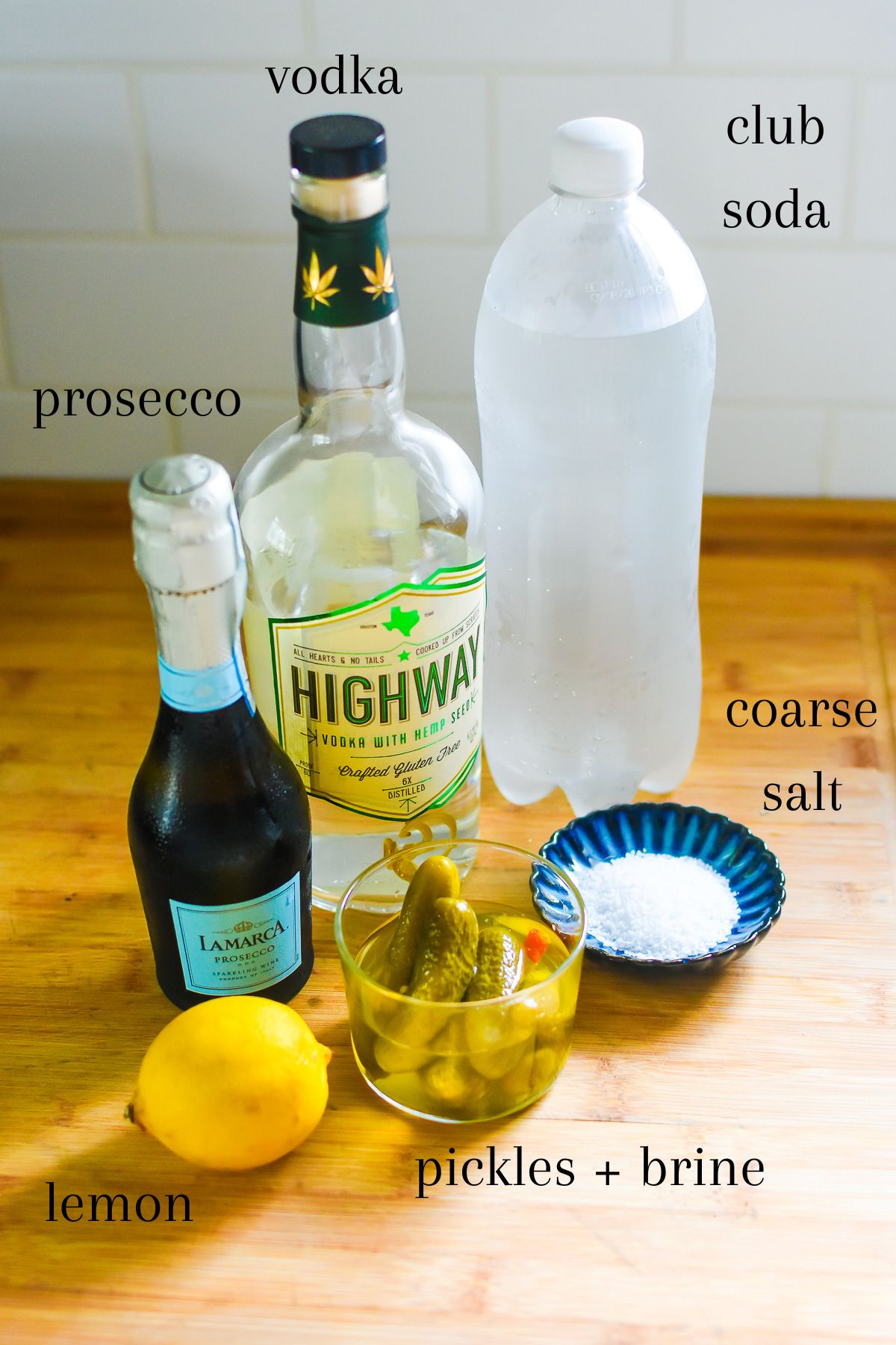 ingredients for making a pickle flavored cocktail on wooden cutting board: bottles of prosecco, vodka, and club soda, pickles in pickle brine, coarse salt, and a fresh lemon.