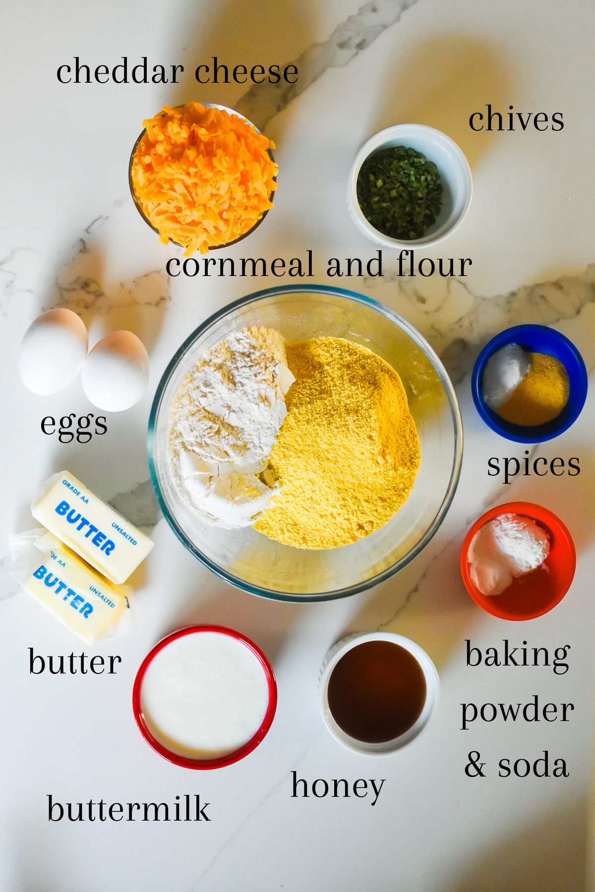 ingredients for cornbread biscuits from scratch on granite counter top: shredded sharp cheddar cheese, freeze-dried chives, eggs, spices, yellow cornmeal, all-purpose flour, unsalted butter, baking powder, baking soda, buttermilk, and honey.