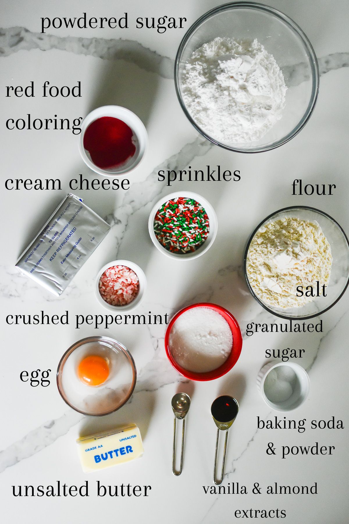 ingredients for a holiday sugar cookies bars recipe on granite counter top: flour, salt, powdered sugar, red food coloring, red and green sprinkles, crushed peppermint candy, cream cheese, granulated sugar, egg, unsalted butter, baking soda, baking powder, vanilla extract, and almond extract.