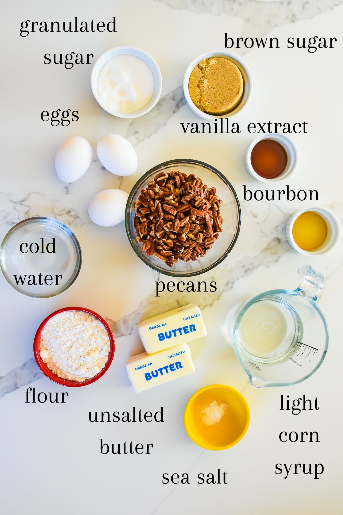 old fashioned ingredients for pecan pie on granite counter top: granulated sugar, brown sugar, eggs, vanilla extract, bourbon whisky, water, pecans, flour, butter, salt, and light corn syrup.
