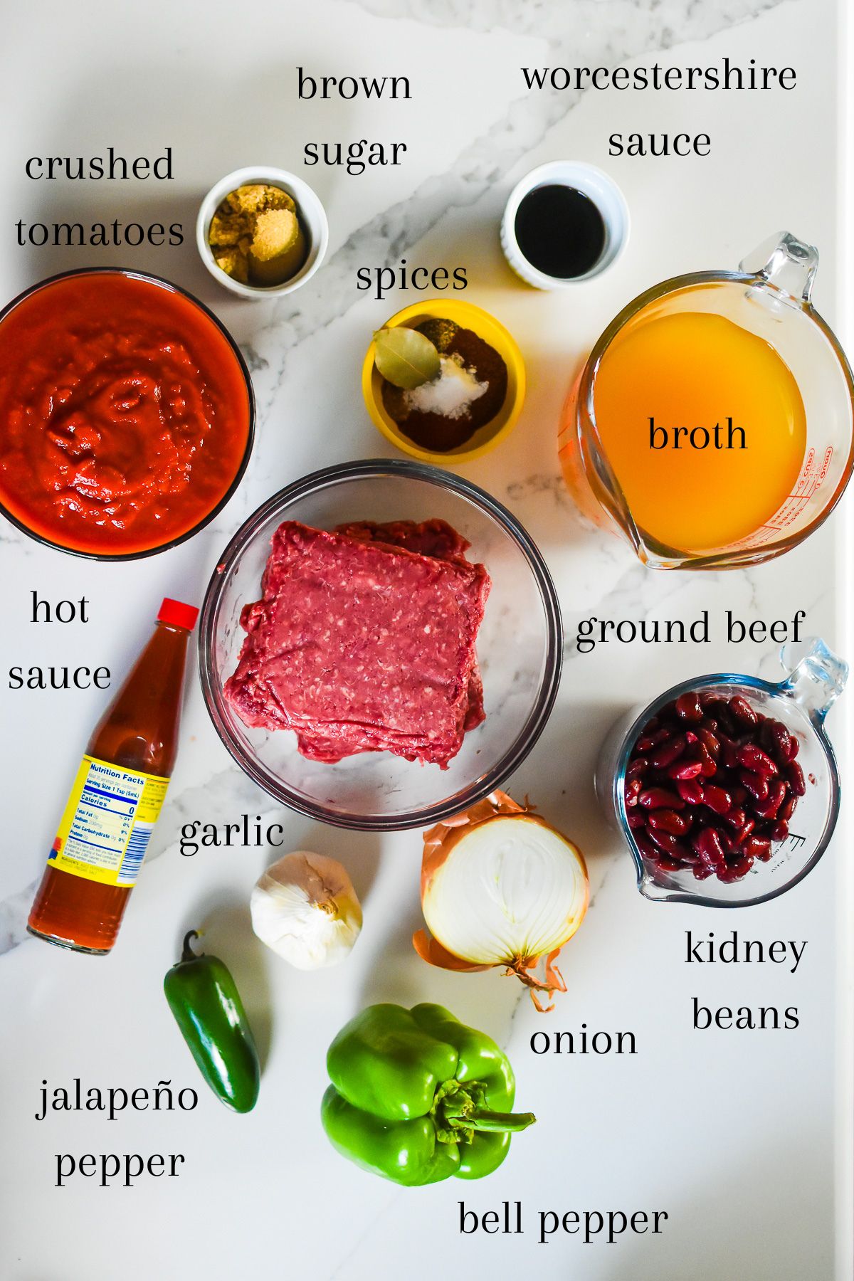 ingredients for making chili using ground beef on granite counter top: crushed tomatoes, brown sugar, Worcestershire sauce, herbs and spices, broth, lean ground beef, hot sauce, dark red kidney beans, onion, garlic, jalapeño, and green bell pepper.
