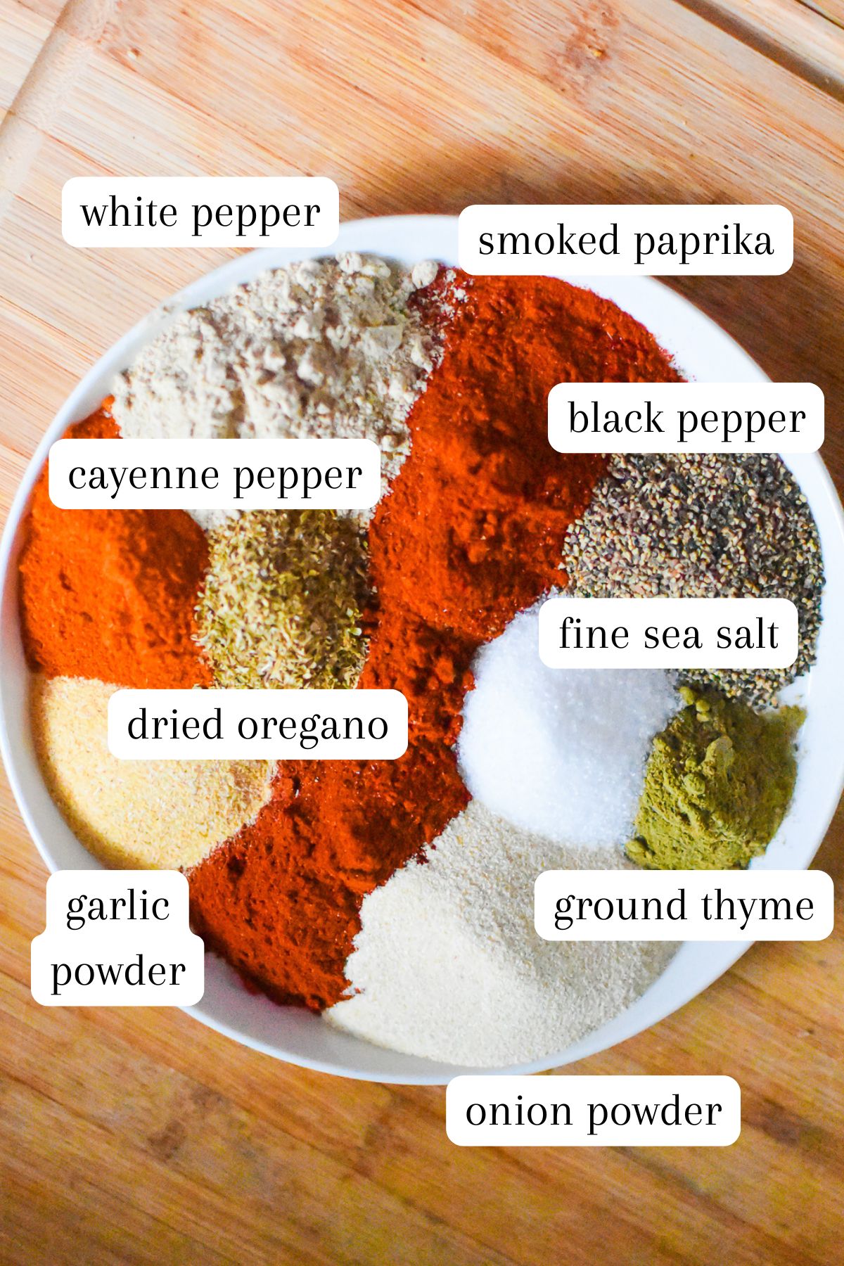 Creole seasoning mix ingredients on white plate: cayenne pepper, white pepper, black pepper, smoked paprika, dried oregano, ground thyme, fine sea salt, garlic powder, and onion powder.
