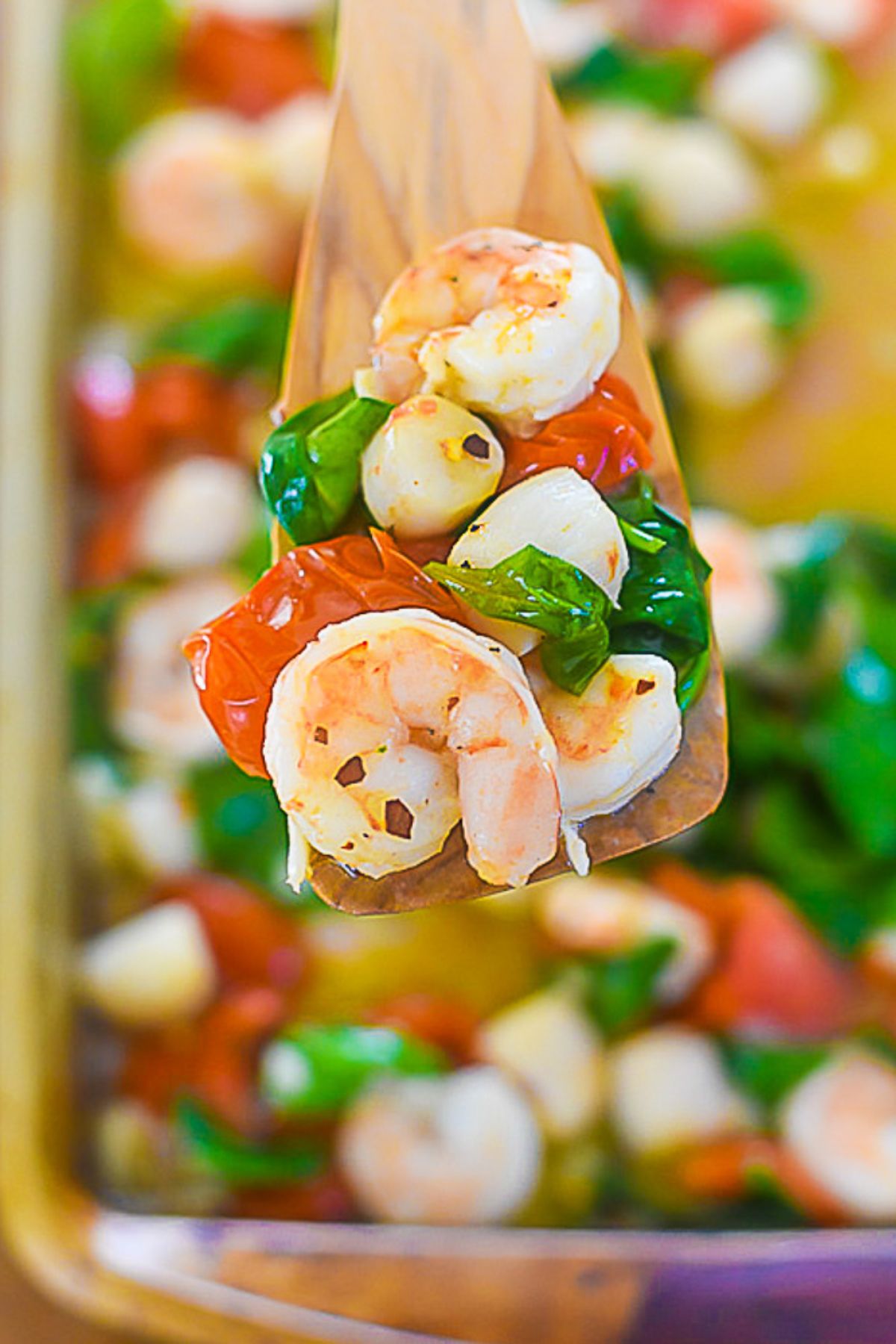 wooden serving spoon serving up scallops, shrimp, tomatoes, and spinach broiled in garlic butter.