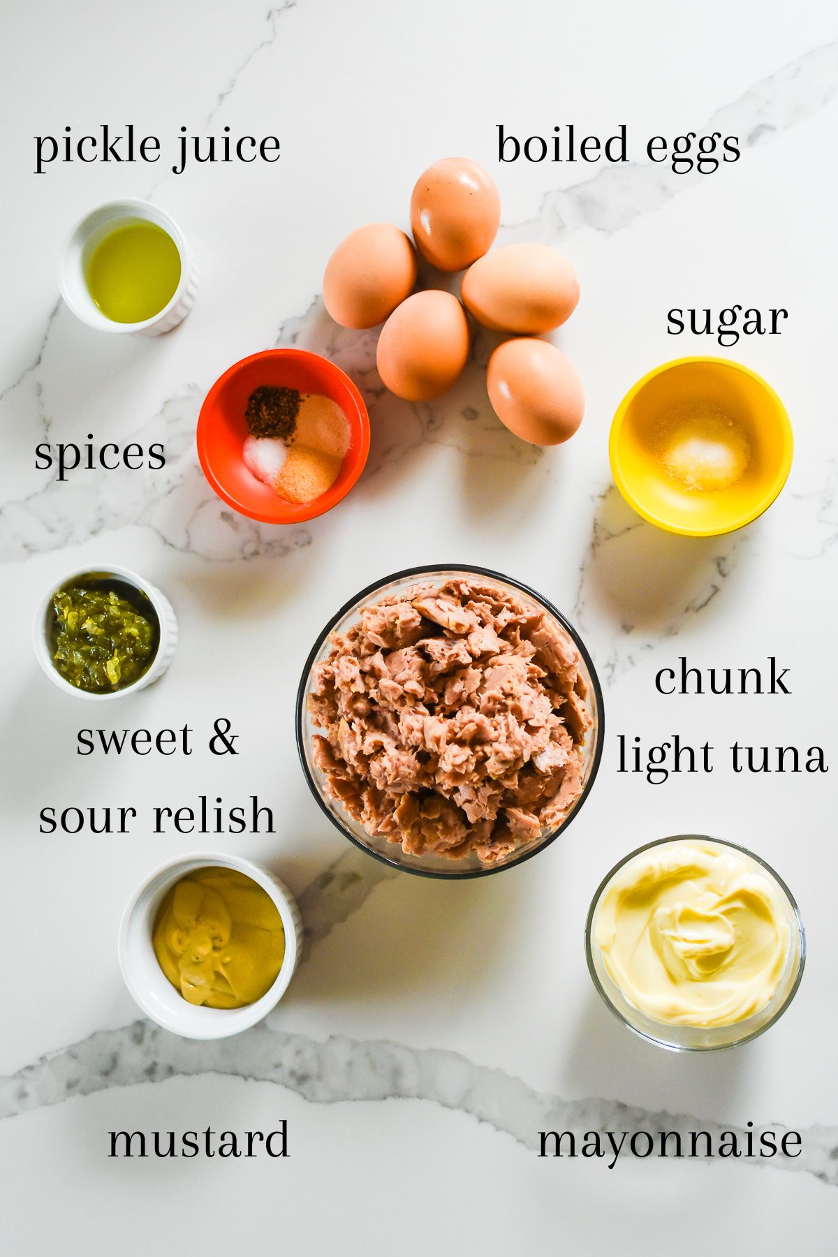 ingredients for a canned tuna fish sandwich recipe on granite counter top: pickle juice, boiled eggs, spices, sugar, chunk light tuna, sweet and sour relish, yellow mustard, and mayo.