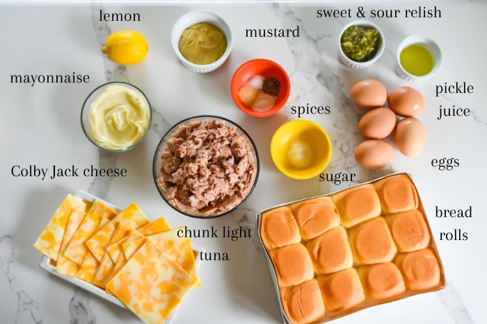 ingredients for making slider-sized baked tuna melts in oven on granite counter top: fresh lemon, mayonnaise, mustard, spices, sugar, relish, pickle juice, boiled eggs, bread rolls, chunk light tuna, and slices of Colby Jack cheese.