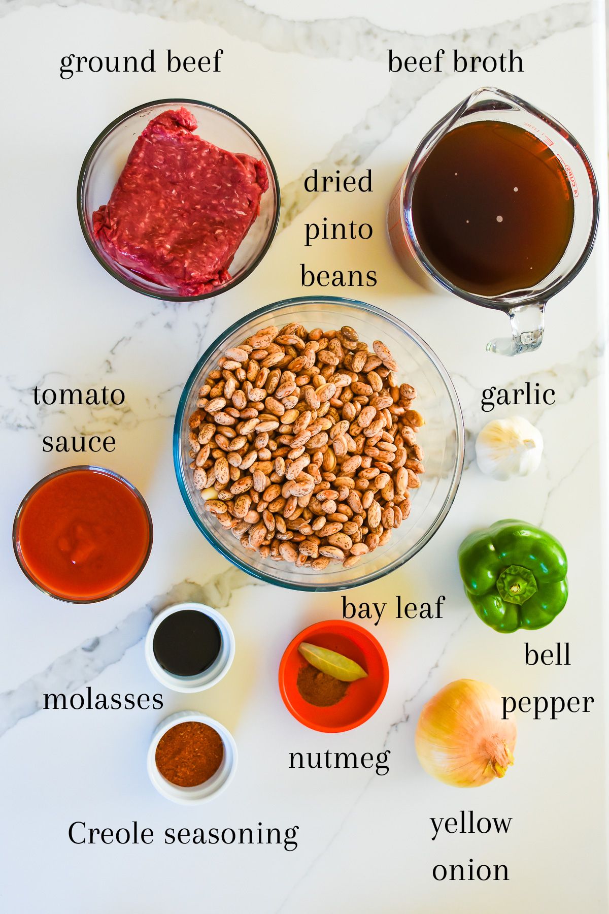 ingredients for making old fashioned pinto beans and ground beef recipe on granite counter top: lean ground beef, beef broth, dried pinto beans, garlic, onion, tomato sauce, bell pepper, molasses, bay leaf, ground nutmeg, and Creole seasoning.