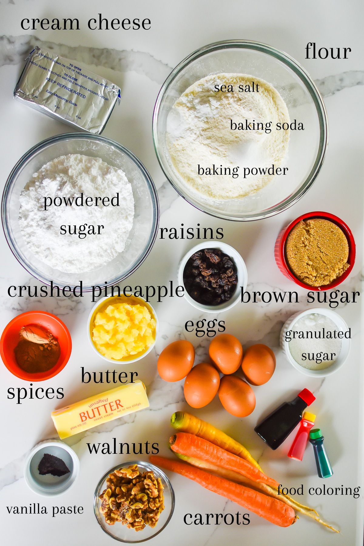ingredients for making a simple carrot sheet cake recipe on granite counter top: block of cream cheese, bowl of flour, sea salt, baking soda, and baking powder, powdered sugar, raisins, brown sugar, granulated sugar, crushed pineapple, butter, eggs, vanilla bean paste, walnuts, carrots, and food coloring.