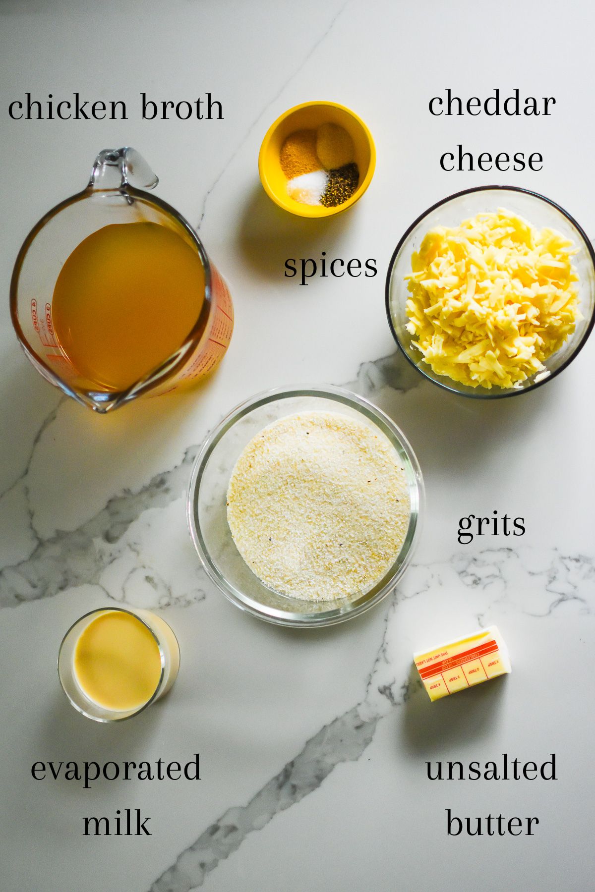 ingredients for the best cheese grits recipe on granite counter top: chicken broth, garlic powder, onion powder, salt, pepper, shredded white cheddar cheese, white grits, evaporated milk, and unsalted butter.