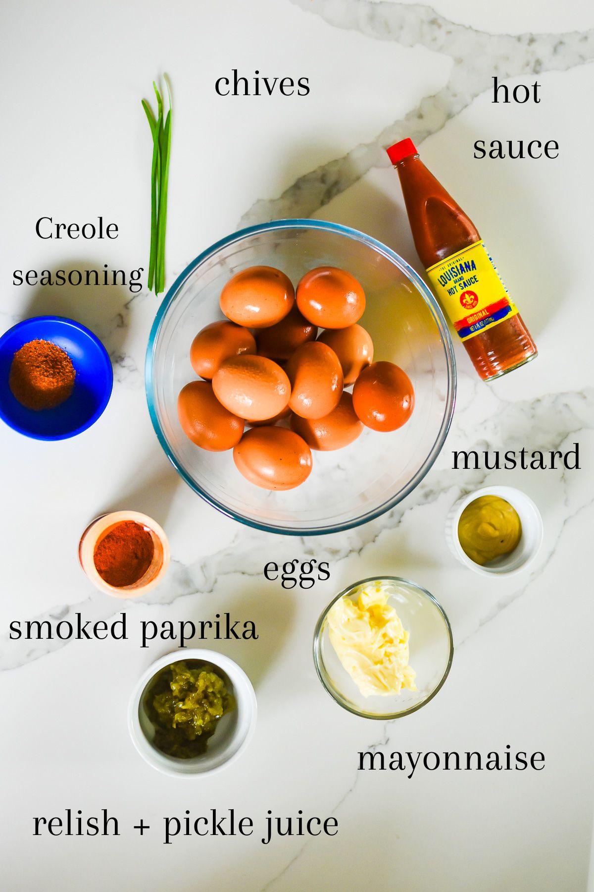 ingredients for old fashioned black southern deviled eggs on granite counter top: fresh garlic chives, hot sauce, Creole Cajun seasoning, eggs, mustard, mayo, pickle relish, and smoked paprika.