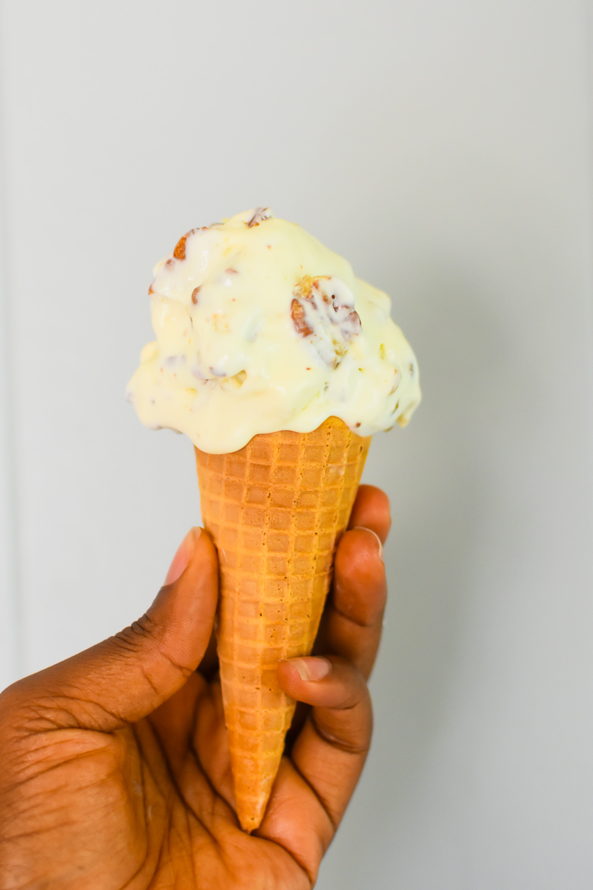 brown hand holding a waffle cone topped with a scoop of creamy, eggless butter pecan ice cream.
