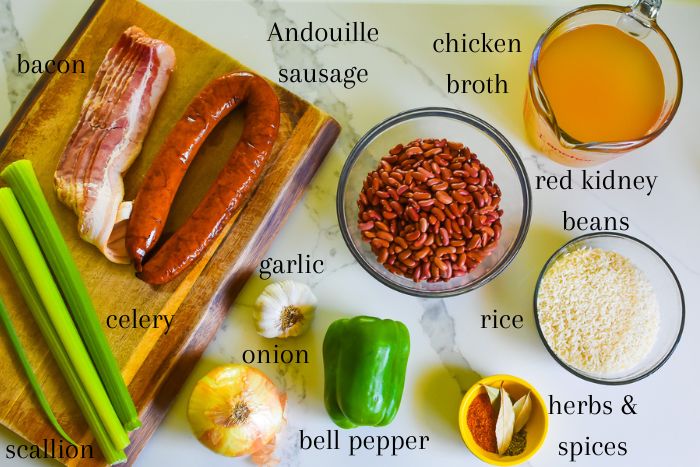 red beans and rice ingredients on granite counter top: fresh scallion, celery stalks, onion, garlic, and green bell pepper, uncooked bacon, smoked Andouille sausage, mixed dried light and dark red kidney beans, chicken broth, white rice, and herbs & spices.