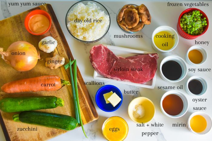 ingredients to make homemade hibachi recipe on kitchen counter top: yum yum sauce, garlic, onion, ginger, carrot, zucchini, scallions, mushrooms, day old white rice, sirloin steak, butter, beaten eggs, salt, white pepper, mirin, sesame oil, oyster sauce, soy sauce, honey, cooking oil, and frozen peas.