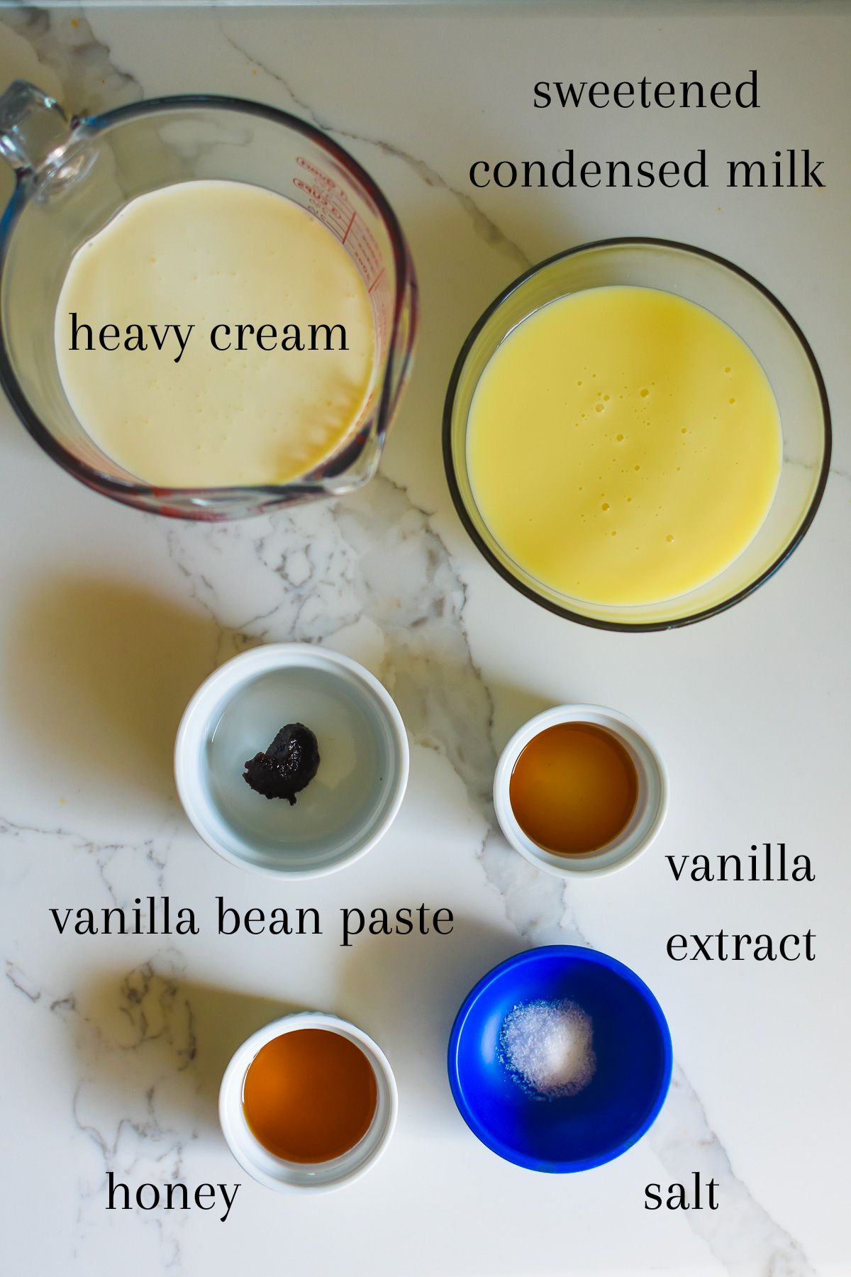 ingredients of homemade vanilla ice cream on granite counter top: heavy cream, sweetened condensed milk, vanilla bean paste, vanilla extract, honey, and sea salt.