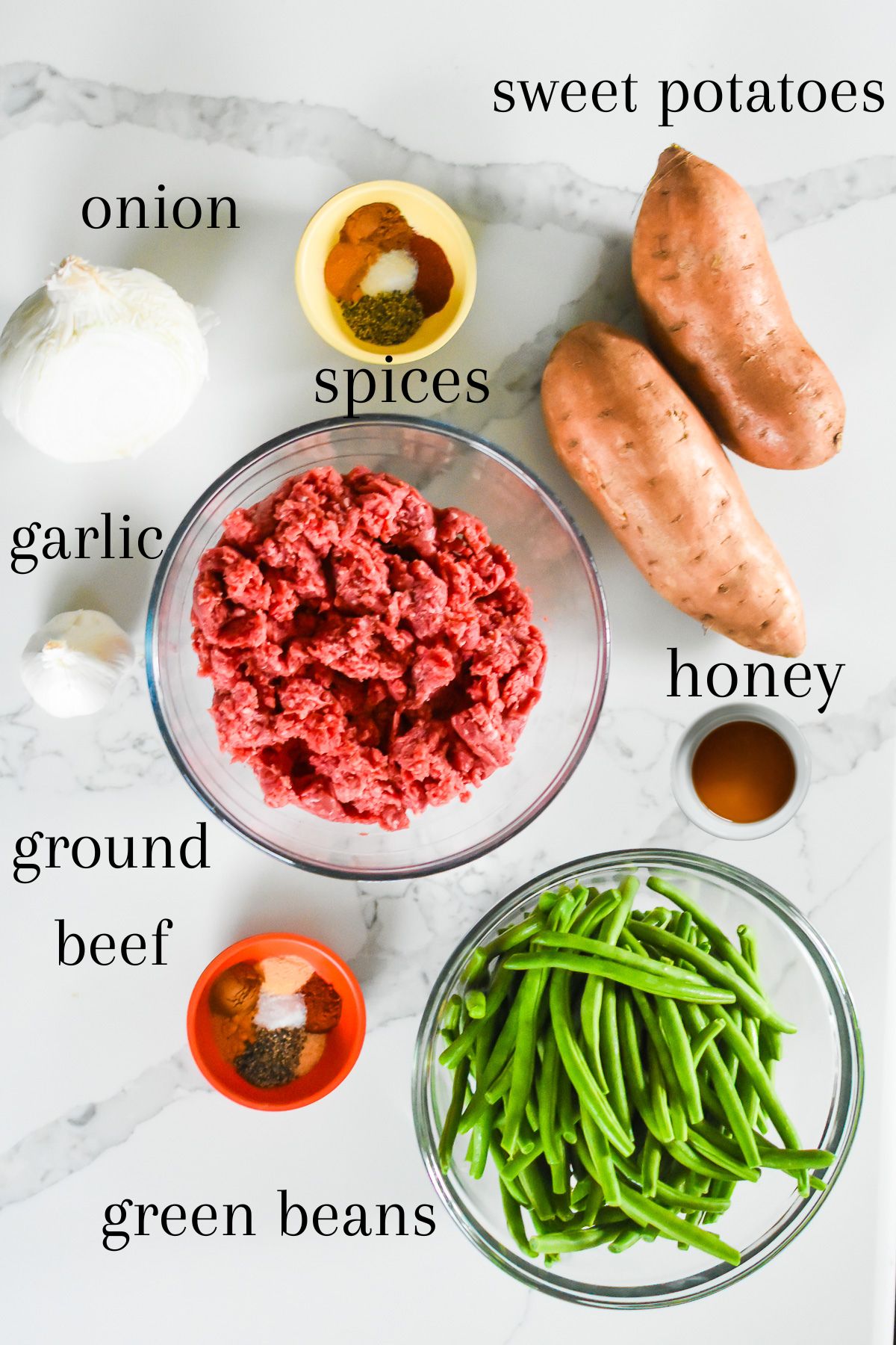 ingredients for making low carb ground beef bowls on kitchen counter top: onion, garlic, spices, sweet potatoes, lean ground beef, honey, and fresh green beans.