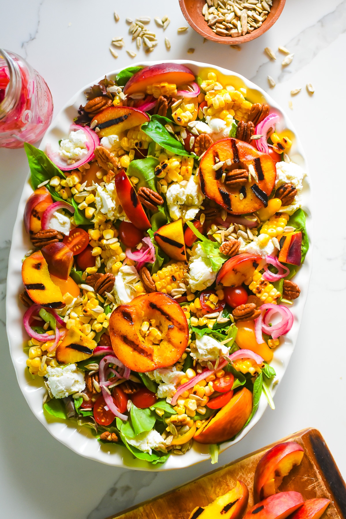 delicious and colorful summer salad with grilled peaches and corn, crunchy nuts and seeds, and goat cheese on a white scalloped platter.