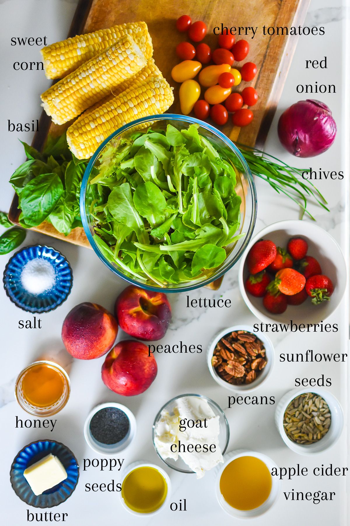 ingredients for an epic summer salad to share on kitchen counter top: ears of yellow corn, cherry tomatoes and yellow pear tomatoes, red onion, chives, basil leaves, butter lettuce, strawberries, peaches, salt, sunflower seeds, pecans, goat cheese, apple cider vinegar, olive oil, poppy seeds, butter, and honey.