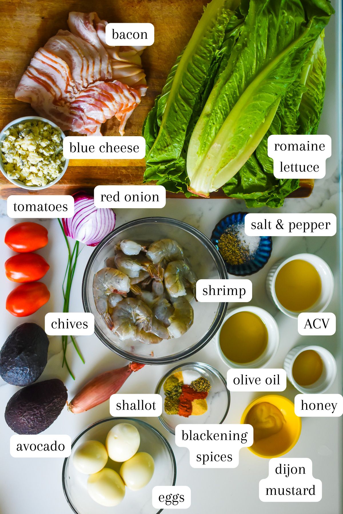 traditional cobb salad recipe ingredients with shrimp on kitchen counter top: bacon, romaine lettuce, blue cheese, red onion, salt and pepper, tomatoes, chives, avocados, shallot, jumbo shrimp, hard-boiled eggs, blackening spices, dijon mustard, olive oil, honey, and apple cider vinegar.
